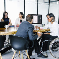 Hispanic transgender businesswoman in wheelchair working on project with group of business people in office, in disability concept and disabled people